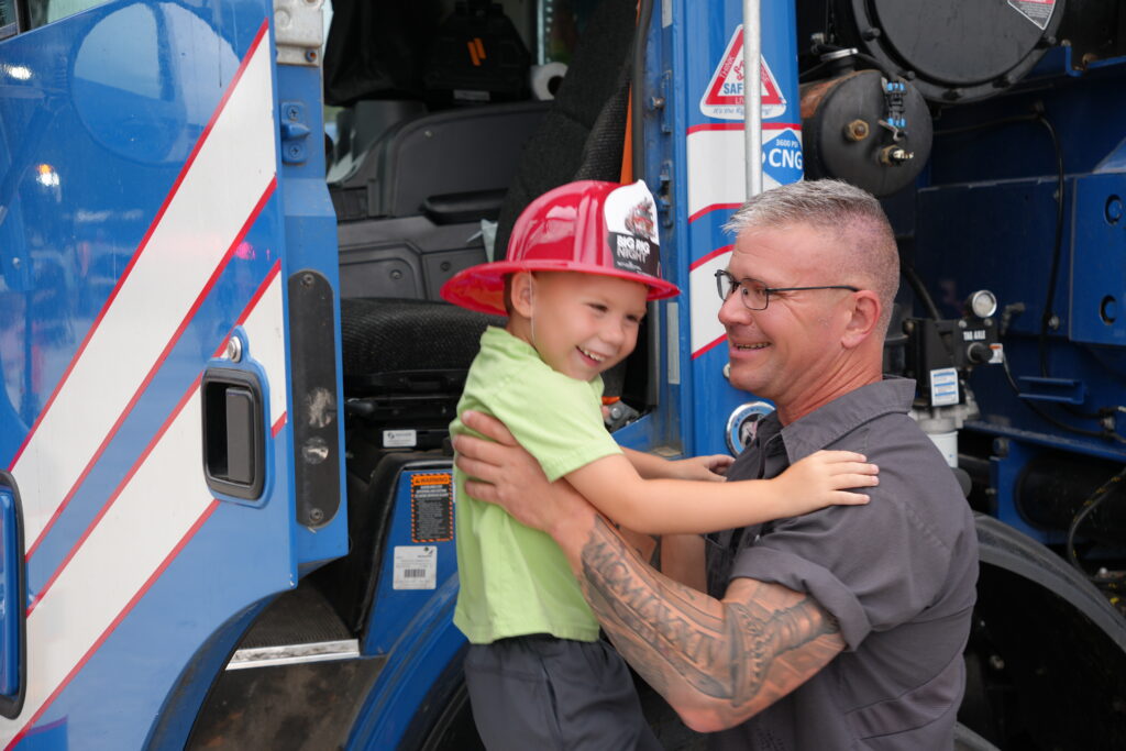 Smiling man helping a child in a firefighter's hat out of the front of an ambulance./